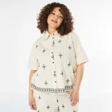 Camisa beige de manga corta con cuello clásico y cierre frontal con botones. Presenta bordados geométricos en color negro.