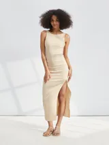 Vestido midi beige ajustado al cuerpo, sin mangas y con escote redondo. Presenta un frunce lateral que genera un drapeado y una abertura alta en la pierna.