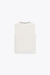 Blusa blanca plisada sin mangas, con cuello redondo y cierre frontal con botones. Presenta vivos negros en cuello, sisas y botonera.