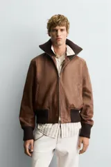 Cazadora de cuero color marrón, estilo bomber, con cuello ancho de punto acanalado marrón oscuro, cierre frontal de cremallera y puños y cintura elásticos de punto acanalado marrón oscuro.