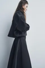 Falda midi negra de gabardina, tiro medio, cintura con trabillas, corte evasé con volumen y cierre frontal con cremallera y botón.
