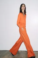 Pantalón ancho color naranja, de tiro medio y cintura con trabillas. Tiene bolsillos laterales y de vivo en espalda, pinzas en delantero, raya planchada y cierre frontal con cremallera, botón y ganchos metálicos.
