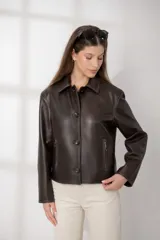 Chaqueta de cuero ovino color marrón oscuro, con cuello camisero, cierre frontal de botones y bolsillos verticales con cierre en la parte delantera. Presenta un corte recto y holgado.