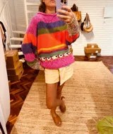 Sweater tejido de lana multicolor con cuello redondo y mangas largas. Tiene rayas horizontales en tonos fucsia, violeta, verde, naranja y amarillo. En la parte inferior tiene un diseño de ovejas blancas sobre fondo fucsia.