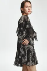 Vestido corto negro con estampado paisley en tonos beige y detalles dorados. Escote en V con lazos ajustables, mangas largas con puños elásticos y volantes en el mismo tejido en el escote y el ruedo.