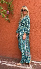 Vestido largo estilo cruzado (wrap dress) de manga larga acampanada, con estampado floral en tonos verde petróleo, amarillo pálido y celeste. Presenta un ribete decorativo en el escote y el borde del cruce.
