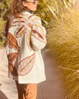 Cárdigan de lana color crudo con apliques de hojas bordadas en tonos marrones, naranjas y amarillos. Corte holgado y largo a la cadera.