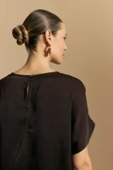 Top negro de manga corta con silueta amplia y caída fluida, confeccionado en un tejido liviano con leve brillo. Presenta un botón de nácar en la espalda con una pequeña abertura y mangas amplias tipo kimono.