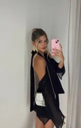 Vestido corto negro con escote pronunciado en V y drapeado en el busto. Presenta mangas largas con aberturas en los hombros (cold shoulder) y puños acampanados.