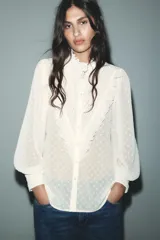 Camisa blanca semitransparente con cuello subido, manga larga con puños, pliegues y volantes combinados. Cierre frontal con botones.