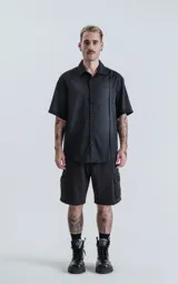 Camisa negra de manga corta con cuello clásico y cierre frontal con botones. Presenta una tira vertical cosida en el frente.