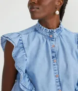 Camisa cropped de jeans celeste, sin mangas, con cuello alto, cierre frontal con botones, volantes en cuello y laterales, y elástico en la cintura.