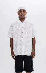 Camisa de manga corta color blanco con textura de toalla y diseño de líneas verticales con círculos. Tiene cuello tipo polo y cierre frontal con botones. 