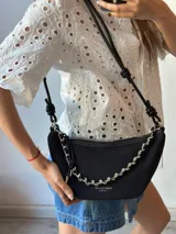 Cartera bandolera negra con cadena plateada decorativa y correa de cuero sintético con nudos.