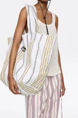 Bolso tipo shopper de lona color crudo con rayas verticales amarillas y grises. Tiene doble asa de hombro y logo de la marca estampado en el frente.