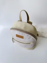 Mochila pequeña de cuero color gris claro y beige, con cierre superior y bolsillo frontal con cierre. Tiene asa de mano y correas ajustables para los hombros.