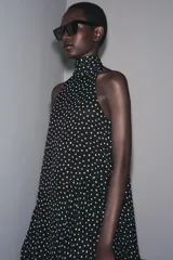 Vestido midi negro con estampado de lunares blancos, cuello halter con pañuelo integrado, sin mangas y bajo asimétrico. Cierre con abertura y botón en la nuca.