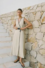 Vestido midi de lino color beige, con escote cruzado en pico y sin mangas. Presenta vivos a contraste en negro y cinturón con hebilla forrada. Cierre frontal con botones.
