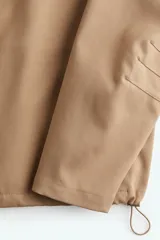 Campera color beige de sarga con cuello, cierre oculto adelante y bolsillos laterales con cierre. Modelo de corte holgado con pinzas en los codos para dar forma y cordón para ajustar elástico con topes en el bajo. Sin forrar.
