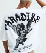 Remera blanca de algodón, corte oversized, con cuello redondo y mangas cortas. Presenta un estampado frontal con un ángel y el texto "PARADISE" en negro, y un pequeño gráfico de estrella/brújula en el lado izquierdo del pecho.