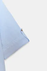 Camisa de lino color celeste claro, con cuello solapa, escote pico, manga corta con vuelta y dos bolsillos de plastrón en el delantero. Cierre frontal con botones.