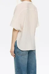 Camisa color beige estilo capa confeccionada en viscosa con textura. Cuenta con cuello camisero con lazo, manga corta murciélago y cierre frontal con botones.