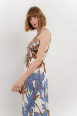 Vestido largo con estampado de hojas en tonos verdes y marrones sobre fondo blanco. Tiene breteles finos, escote en V y detalle de lazo en el frente. La falda es amplia y con vuelo.