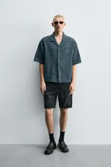 Camisa de denim de algodón, color beige claro, con corte relajado, cuello bowling y manga corta. Presenta cierre frontal con botones a presión y un pequeño bordado a tono en el pecho.