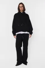 Buzo hoodie de frisa negro, de silueta oversize, con puños y cintura de rib al tono, capucha forrada en su interior y bolsillo canguro. Cordones con ojalillos y punteras metálicas personalizadas con marca. Detalle de tanca para regular cordones. Remaches en boca de bolsillo. Bordado en espalda a contratono.