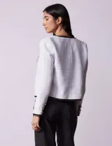 Chaqueta de tweed blanca con ribete negro, escote a la base, abertura central con botones, bolsillos delanteros y puños con aplique de botones perlados.