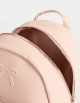 Mochila pequeña de cuerina color rosa pálido, con moño en relieve en el frente y llavero con forma de corazón trenzado. Tiene correas regulables, manija superior, cierre superior, bolsillo frontal con cierre y bolsillos laterales.