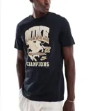 Remera negra de manga corta con estampado frontal en tonos dorados y beige, que incluye el logo "NIKE", un caballo alado, el número "72" y la palabra "CHAMPIONS".