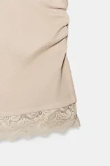 Camiseta ajustada (slim fit) de cuello redondo y manga sisa, color beige claro, con frunces laterales y bajo asimétrico con terminación de encaje a tono.