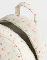 Mochila pequeña de cuerina color crema con estampado de flores pequeñas en tonos rosa, naranja y celeste. Tiene un bolsillo frontal con cierre, correas regulables, manija superior, cierre superior y bolsillos laterales.