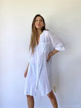 Vestido camisero blanco de corte holgado, con cuello mao, cierre frontal con botones, mangas 3/4 con puños abotonados, dos bolsillos de parche en el pecho y aberturas laterales en el ruedo.