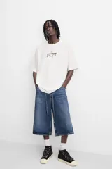 Camiseta blanca de corte relajado, cuello redondo y manga corta. Presenta un estampado de gráficos y textos combinados a contraste en la parte delantera y trasera.