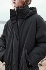 Anorak largo negro resistente al agua y al viento, con aislamiento térmico, cuello alto con capucha ajustable, bolsillos con cremallera, acolchado interior, tirantes elásticos y cremalleras laterales en el bajo.