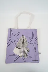 Bolso tote de tela color lila con asas de tela cruda. Presenta un estampado frontal de una figura femenina con abrigo de piel, pantalón a cuadros y lentes de sol, rodeada de estrellas. Incluye el logo de la marca "INDIAN" en la parte inferior.