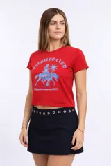 Remera roja de manga corta y calce ajustado (baby tee) con cuello redondo. Presenta un estampado frontal en celeste que incluye el texto "DELOALTO CLUB", una figura femenina con cola de sirena montando a caballo, una palmera y el texto "Deloalto mvdeo est. 2015".
