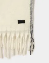 Bufanda tejida color crema con flecos en los extremos y etiqueta de cuero sintético negra.