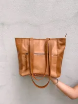 Bolso tipo tote de cuero color camel con paneles laterales texturizados. Tiene doble asa de hombro, cierre superior con cremallera y bolsillos internos.