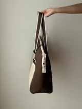 Bolso tipo tote de neopreno color negro y marrón, con cierre superior, bolsillo frontal marrón con logo rojo, bolsillo interno y externo con cierre, doble asa de mano marrón con detalles blancos y opción de correa larga.