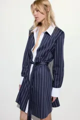 Vestido corto estilo camisero a rayas verticales azul marino y blancas, con cuello y puños blancos. Tiene cierre frontal con botones y lazo para atar en la cintura.