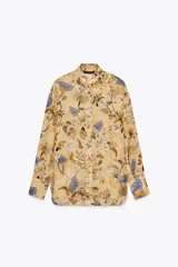 Camisa de ramio color beige con estampado floral en tonos marrones y azules, cuello solapa, manga larga con puños abotonados, bajo asimétrico con aberturas laterales y cierre frontal con botones.