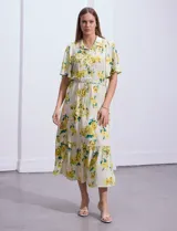 Vestido largo camisero con estampado floral amarillo y verde sobre fondo blanco. Tiene cuello camisero, mangas cortas con volados, cintura con lazo ajustable y falda escalonada con volados.