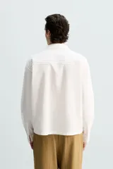 Camisa blanca de corte relajado confeccionada en tejido de algodón y lino. Tiene cuello bowling, manga larga con puños abotonados, jaretas en el delantero, bolsillos de parche en pecho y cadera, bajo con aberturas laterales y cierre frontal con botones.