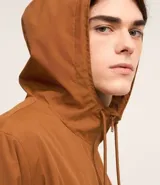 Campera rompevientos color verde oliva, con capucha ajustable con cordón, cierre frontal con cremallera, bolsillos laterales con cremallera y puños elásticos.