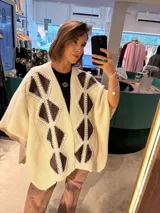 Ruana color crudo de tejido de punto grueso, con cuello alto y abertura frontal. Presenta detalles de rombos tejidos en color ladrillo y negro en la parte delantera.