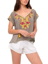 Blusa color taupe de mangas cortas con bordados florales en amarillo y fucsia. Tiene escote redondo con lazo y borlas amarillas.