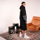 Sudadera tipo canguro negra con capucha y bolsillo canguro frontal. Presenta un diseño de doble manga, donde las mangas interiores son de color gris oscuro. Tiene un pequeño logo bordado en blanco en el pecho izquierdo.